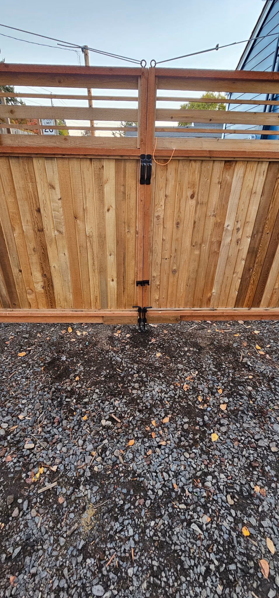 Double wooden gate with black metal latches on a gravel driveway under clear sky.