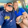 Man wearing sunglasses and a navy shirt sitting next to a woman in a blue dress inside a car.