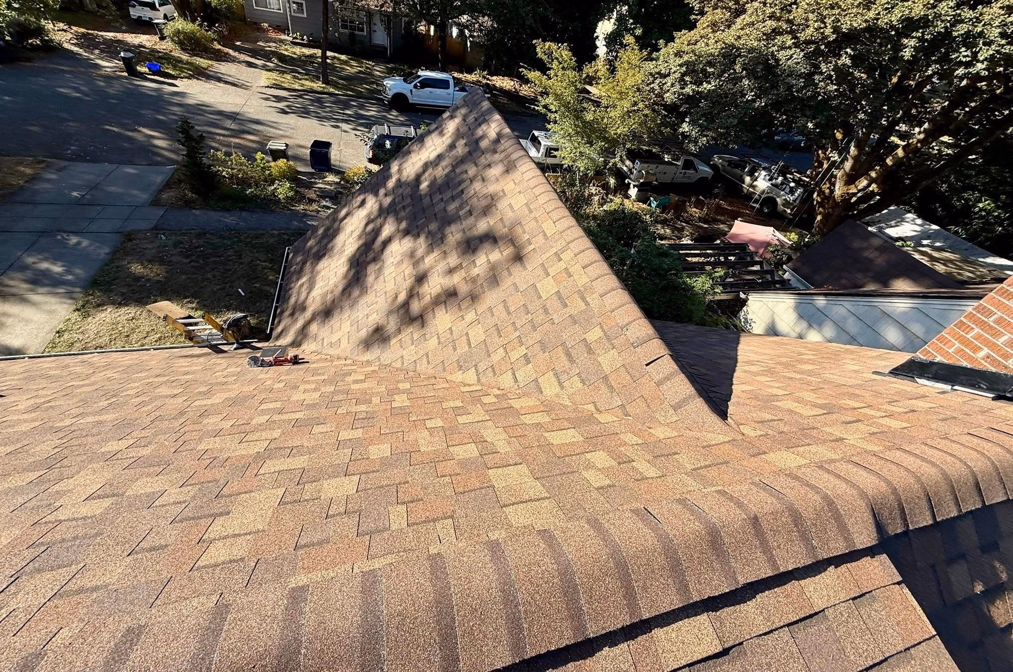 View of a newly shingled brown roof from the top, with shadows of trees and surrounding yard and street below.
